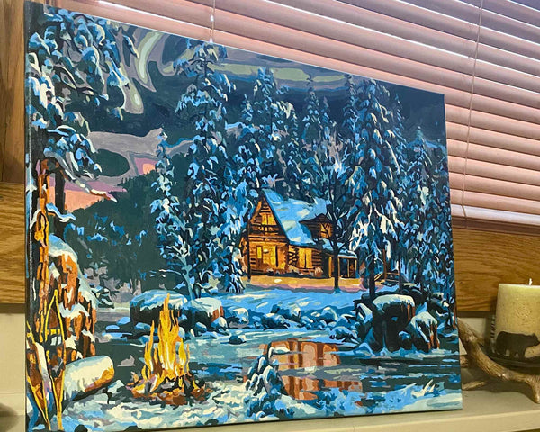 Aurora Bliss paint by numbers completed by Tammie S: a lit cabin in snowy pines beside a campfire under the northern lights